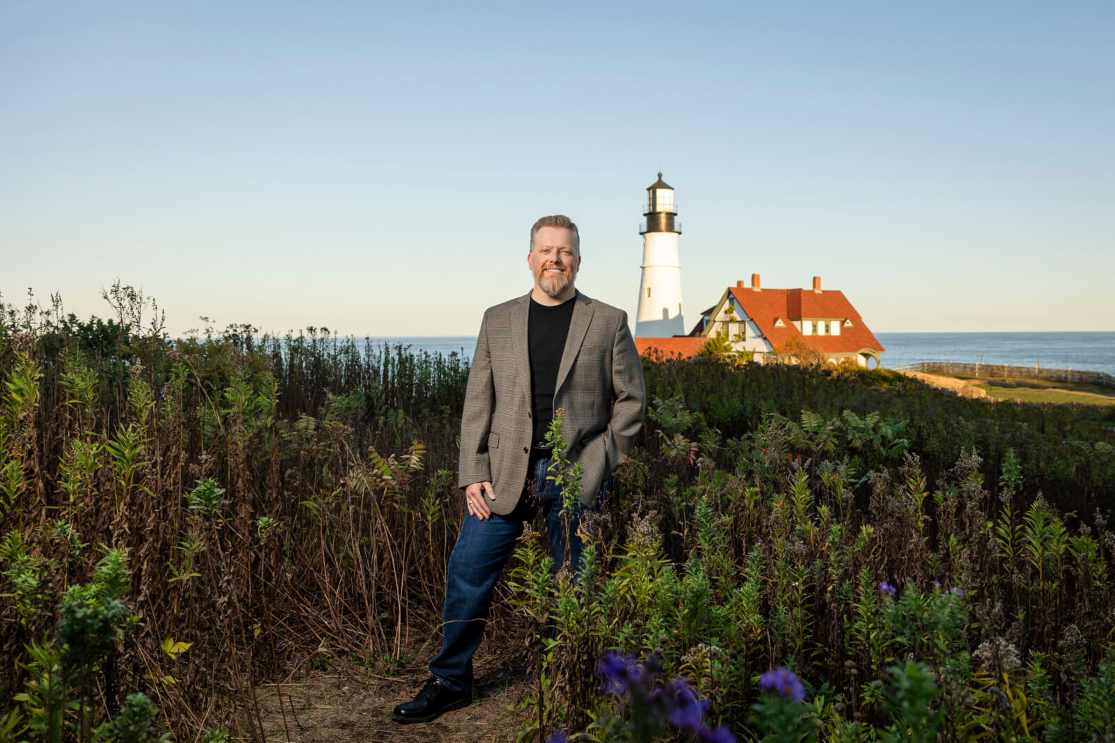 Boots2Roots Executive Director Randy Bell stands among tall grass with a charming lighthouse and water in the background.