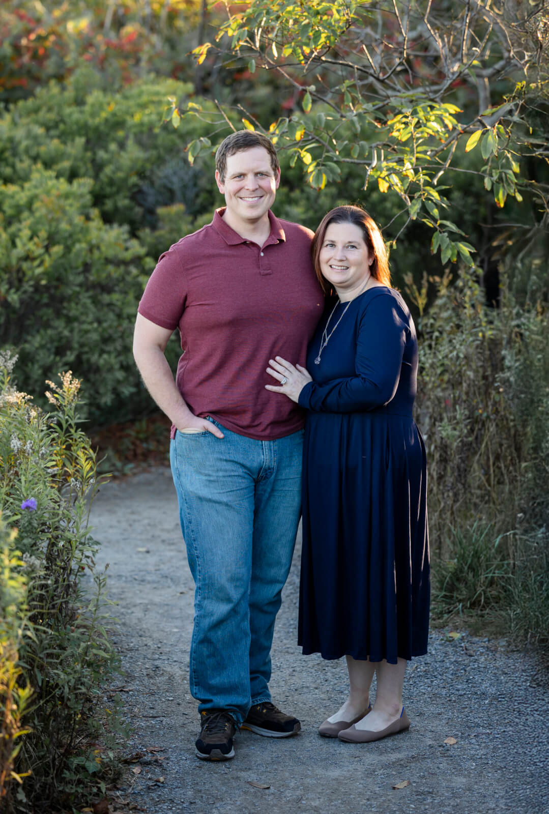 A couple stands on a gravel path surrounded by trees, tall grass and flowers.
