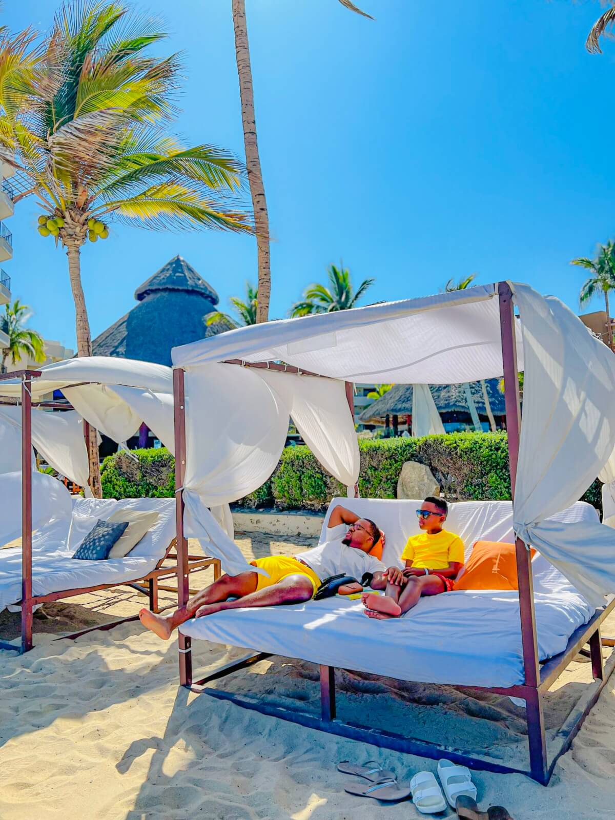 Military child and father sitting on white bed under cabana on sand and surrounded by palm trees while on vacation in Mexico. 