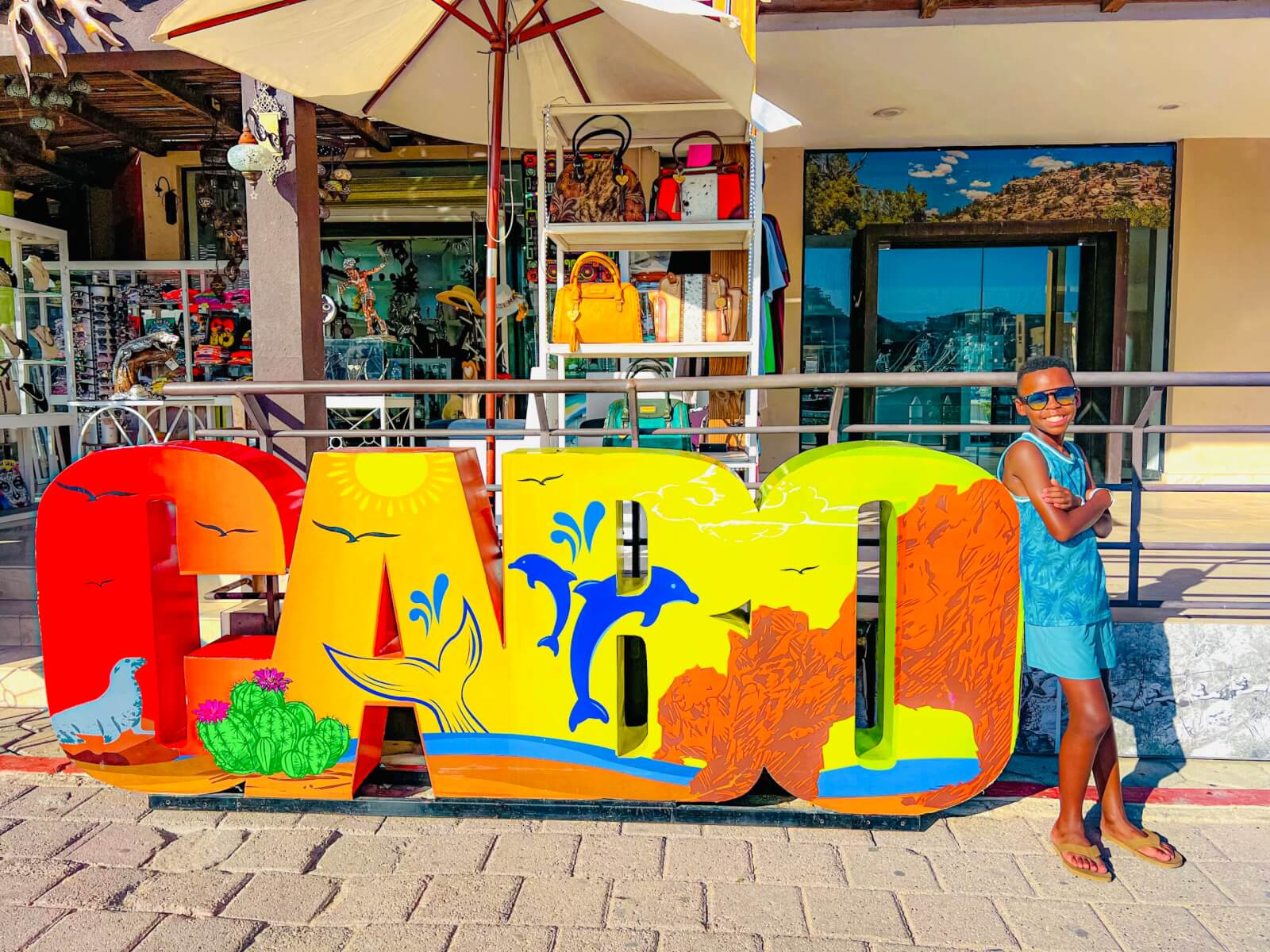 Military child smiling in his sunglasses as he leans against a brightly colored "CABO" sign on vacation in Mexico.