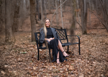 Ashlee Sullivan of WOMAN + WARRIOR sits outside on a black bench surrounded by fallen leaves.