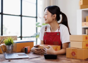 Female entrepreneur wearing red apron smiles as she sits at her desk in front of large window with packages all around her.