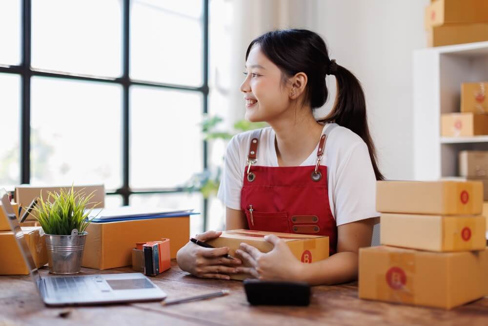 Female entrepreneur wearing red apron smiles as she sits at her desk in front of large window with packages all around her.