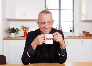 Actor Tom Hanks wears black in a white kitchen while holding a white coffee mug with HANKS across it.