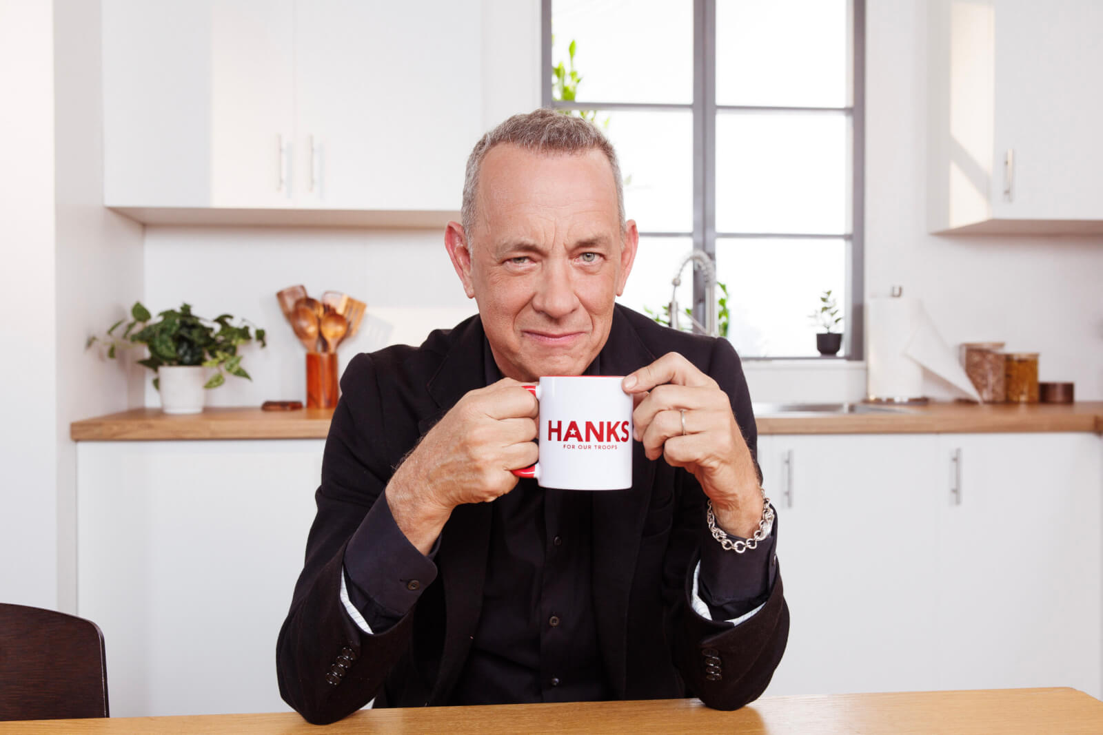 Actor Tom Hanks wears black in a white kitchen while holding a white coffee mug with HANKS across it.