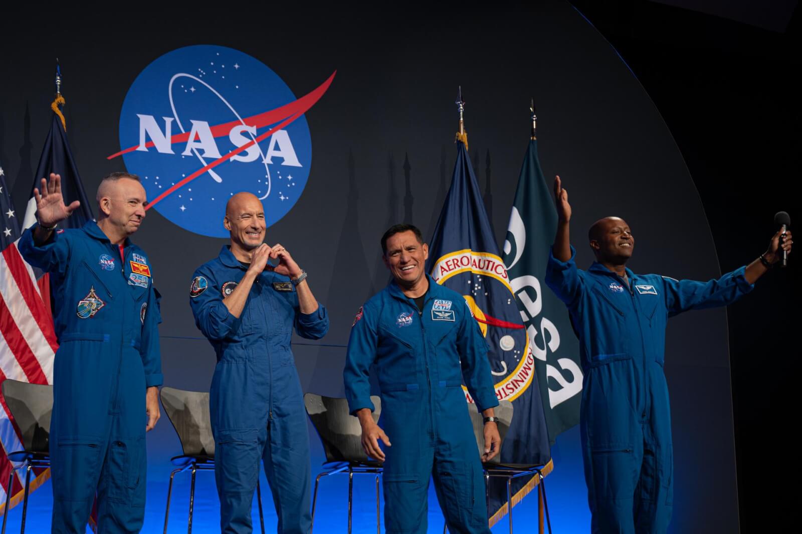 The four Artemis III crew members smile in their blue astronaut suits, with flags and the NASA logo behind them. 