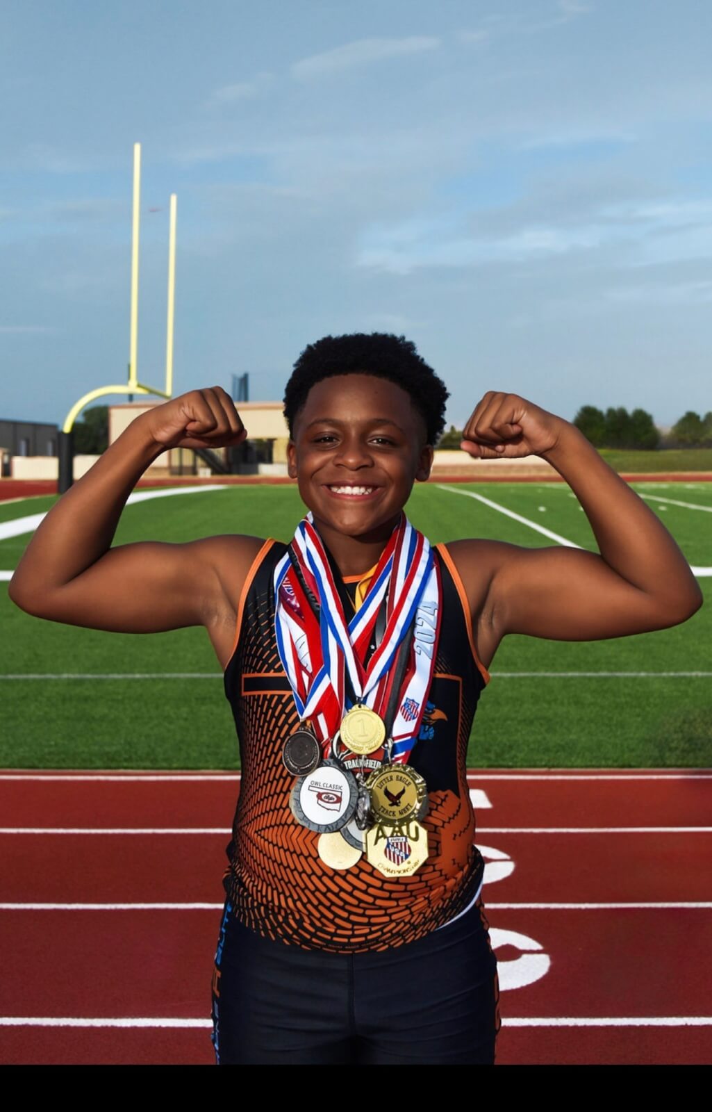 Military kid Khai Pollard stands on a track with medals around his neck, and shows off his muscles while smiling.